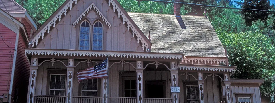 The Lace House in Black Hawk: a small Carpenter Gothic house with lacy white trim along its steep gable and porch, a flag on the porch and pines on the slope behind