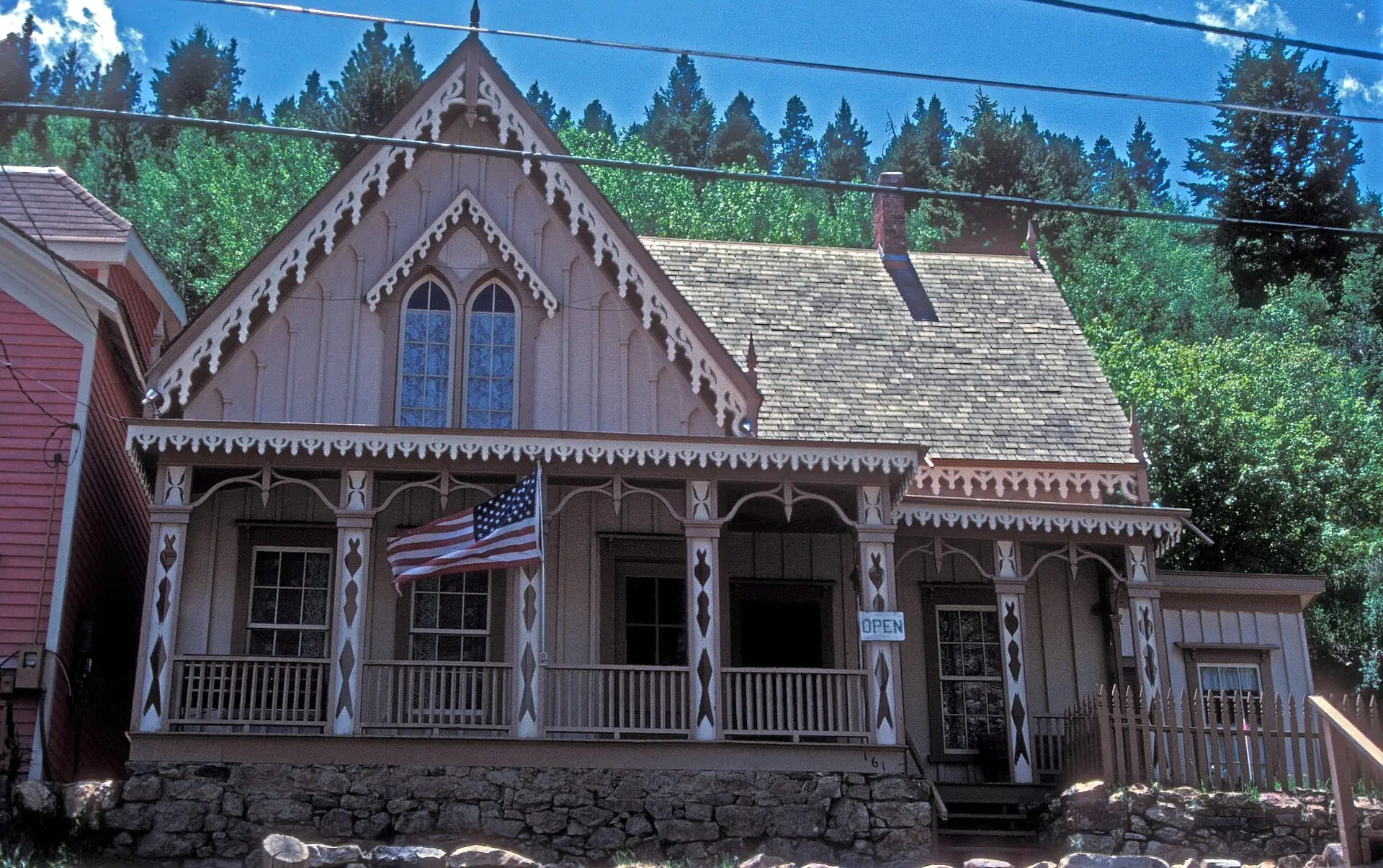 The Lace House in Black Hawk: a small Carpenter Gothic house with lacy white trim along its steep gable and porch, a flag on the porch and pines on the slope behind